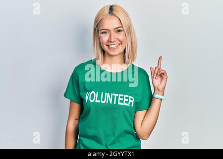 Belle femme blonde portant le t-shirt volontaire gai avec un sourire sur le visage pointant avec la main et le doigt vers le haut sur le côté avec l'exp heureux et naturel Banque D'Images