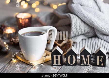 Composition hivernale confortable avec une tasse de thé, le mot décoratif Home, des éléments tricotés et des lumières bokeh. Banque D'Images