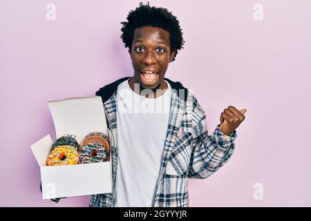 Jeune afro-américain tenant une boîte de beignets colorés savoureuse pointant le pouce vers le côté souriant heureux avec la bouche ouverte Banque D'Images