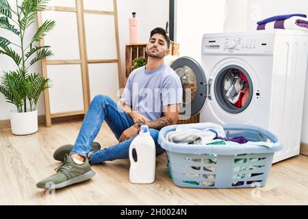 Jeune homme hispanique mettant le linge sale dans le lave-linge détendu avec une expression sérieuse sur le visage. Simple et naturel regarder la caméra. Banque D'Images