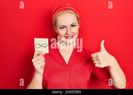 Jeune femme blonde tenant le symbole infini rappel souriant heureux et positif, pouce vers le haut faisant excellent et signe d'approbation Banque D'Images