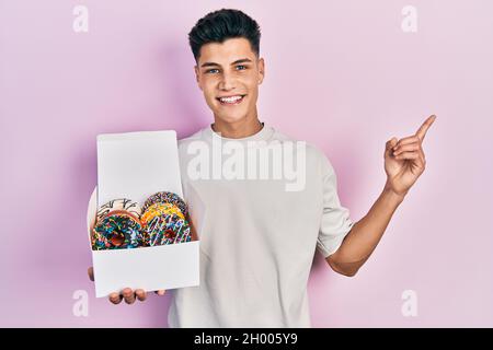 Jeune homme hispanique tenant une boîte de beignets colorés et savoureux souriant, souriant, avec la main et le doigt sur le côté Banque D'Images