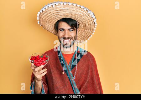 Un jeune hispanique portant un chapeau mexicain tenant un Chili qui a l'air positif et heureux debout et souriant avec un sourire confiant montrant des dents Banque D'Images