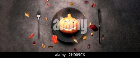 Citrouille avec des yeux brûlants pour Halloween sur une assiette noire avec une fourchette et une cuillère. Chute des feuilles d'automne. Vacances effrayantes, horreur, photo panoramique. Banque D'Images