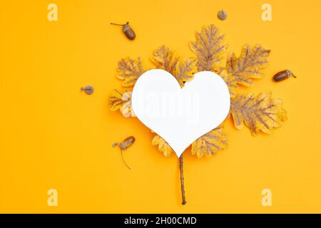 Arrière-plan d'automne vibrant composé de feuilles d'automne pigmentées, d'acornes et d'un papier en forme de cœur découpé sur un fond jaune. Banque D'Images