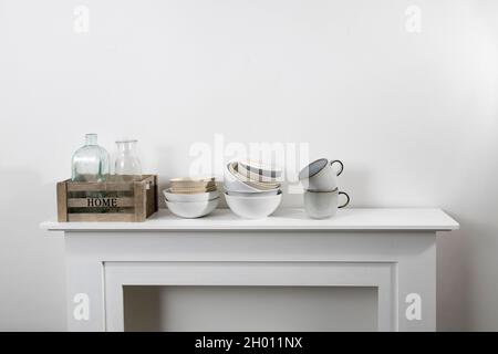 les ustensiles de cuisine, les tasses de différentes tailles et nuances, les bols, les bouteilles, les articles de rangement, sont sur une commode blanche. Banque D'Images