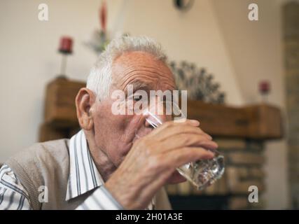 Un vieil homme boit de l'eau du verre à la table à la maison.Concept d'hydratation chez les personnes âgées Banque D'Images