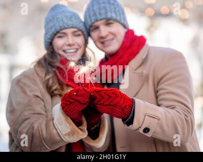 Jeunes fêtes joyeux homme et jolie femme en tenues d'hiver tenant des lumières bengale et souriant à l'appareil photo, heureux couple célébrant des holi d'hiver Banque D'Images