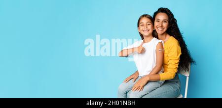 Vaccination de l'enfant.Joyeux Moyen-Orient mère et fille montrant bras vacciné et comme geste approuvant vaccin contre Covid-19 sur Blue B. Banque D'Images