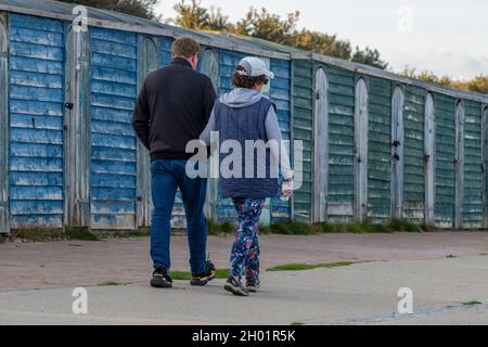 un couple âgé d'âge moyen marchant bras dans le bras le long d'un front de mer avec des huttes de plage de vieux chemin de fer peintes en bleu à l'arrière-plan sur l'île de wight. Banque D'Images