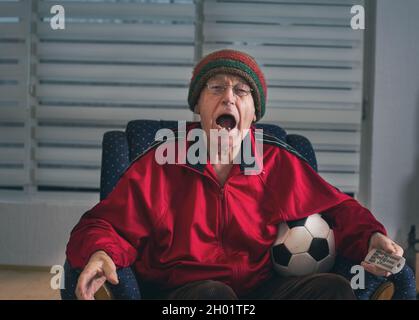 Un homme senior enthousiaste assis dans un fauteuil à la maison, tenant le ballon de football et regardant un match de football à la télévision Banque D'Images