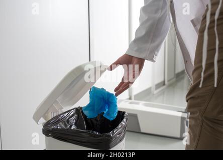 Gros plan de la main du médecin en manteau blanc jeter des couvre-chaussures de protection à usage unique dans une poubelle Banque D'Images