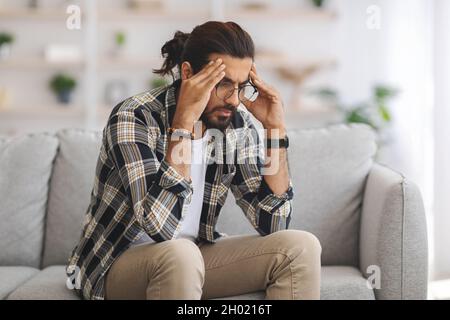 Homme indien barbu dans une ambiance décontractée avec des lunettes assises sur un canapé seul à la maison, touchant sa tête et regardant vers le bas, souffrant de maux de tête ou de l'expérience Banque D'Images