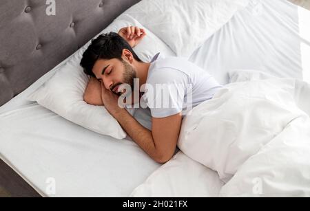 Jeune arabe endormi, reposant paisiblement dans un lit confortable, allongé avec des yeux fermés, espace libre.Loisirs, sommeil profond masculin, temps de repos a Banque D'Images