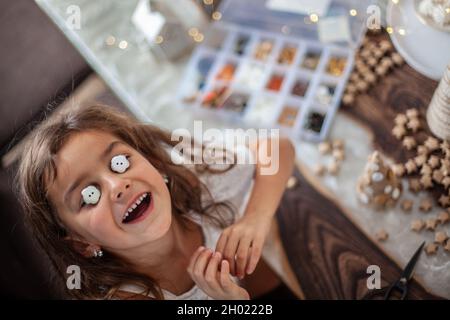 Jolie petite fille avec cheveux bouclés faire des métiers et décorer le cône de sapin de Noël avec des boutons. Banque D'Images