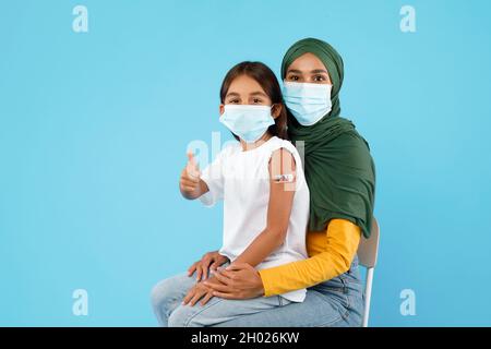 Vaccination des enfants contre le coronavirus.La mère et la fille islamiques vaccinées gesturant des Thumbs vers le haut montrant le bras avec le plâtre portant le visage masques au-dessus de Blue Stu Banque D'Images