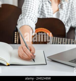 Femme écrivant une chanson, prenant des notes dans un bloc-notes.Enseignement de la guitare musicale en ligne.Cours de formation en ligne jouant de la guitare à la maison.Mains de femmes Banque D'Images