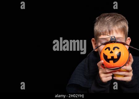 Un enfant tenant un panier orange en forme de citrouille avec une face à manger, une lanterne de Jack et une chauve-souris sur un fond noir. Le garçon attend Halloween Banque D'Images