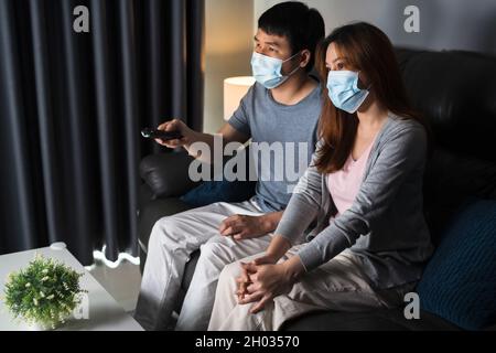 Jeune couple regardant la télévision sur un canapé et portant un masque médical pour protéger le coronavirus (Covid-19) Banque D'Images