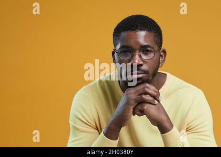 Portrait minimal d'un homme afro-américain portant des lunettes et regardant l'appareil photo tout en posant le menton à la main et en posant sur fond jaune, espace de copie Banque D'Images