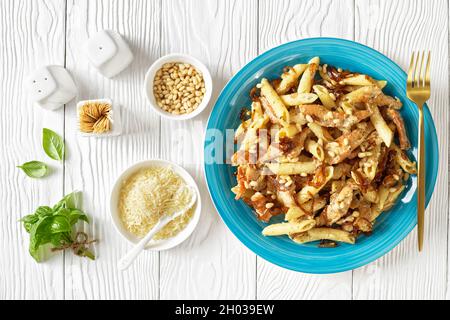 penne de pâtes alfredo de poulet à l'oignon caramélisé arrosé de pignons de pin et de parmesan râpé sur une assiette bleue sur une table en bois blanc, horizont Banque D'Images