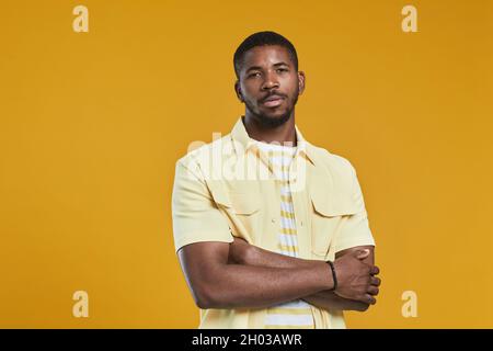 Portrait à la taille de l'homme afro-américain beau regardant l'appareil photo tout en posant en toute confiance avec les bras croisés sur fond jaune dans le studio, espace de copie Banque D'Images