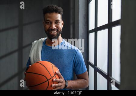 Joyeux jeune joueur de basket-ball afro-américain debout à l'intérieur à la salle de gym, regardant l'appareil photo. Banque D'Images