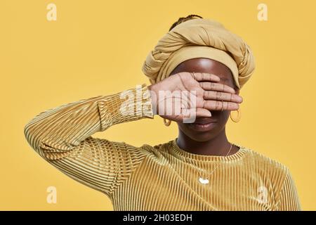 Portrait minimal d'une jeune femme afro-américaine portant un couvre-tête et couvrant les yeux tout en se tenant sur fond jaune dans un studio, espace de copie Banque D'Images