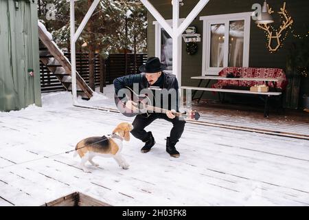 Musicien excentrique dans un chapeau et une veste en cuir jouant de la guitare pour un chien sympathique.Il est debout devant la maison sur un plancher de planche enneigée. Banque D'Images