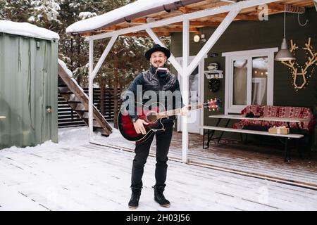 Musicien dans un chapeau et une veste de cuir debout devant la maison sur un plancher de planche enneigée.Il a de la guitare et de l'harmonica. Banque D'Images