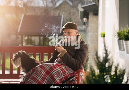 Homme senior assis sur un banc avec un chien dans le jardin recouvert d'une couverture et d'un tuyau de fumée Banque D'Images