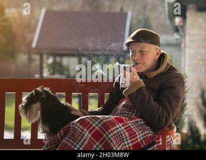 Homme senior assis sur un banc avec un chien dans le jardin recouvert d'une couverture et d'un tuyau de fumée Banque D'Images