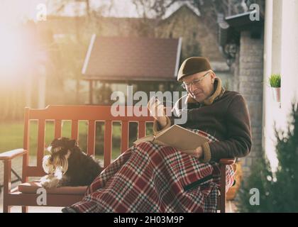 Homme senior assis sur une banque dans le jardin couvert d'un livre de lecture de couverture, d'une pipe à fumer et d'un chien en cuddling Banque D'Images