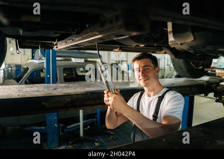 Portrait moyen de beau professionnel souriant mécanicien de voiture en uniforme debout dans la fosse d'inspection et de travail avec l'outil Banque D'Images