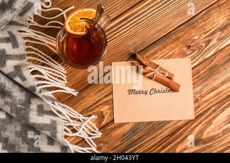 vue de dessus d'une tasse de thé avec des tranches d'orange près des bâtons de cannelle sur la carte de voeux avec du merry christmas lettering sur une surface en bois Banque D'Images