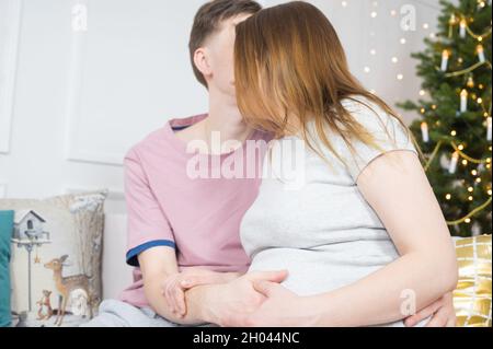 Heureux mari embrassant et prenant soin de sa femme enceinte sur le canapé, fond de Noël Banque D'Images