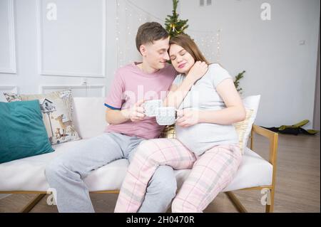 Mari prenant soin de sa femme enceinte, embrassant et embrassant. Un jeune couple se détende sur un canapé à l'heure de Noël Banque D'Images
