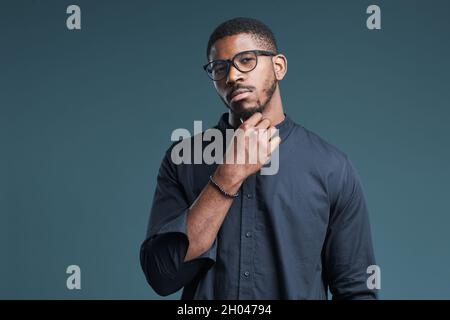 Portrait à la taille de l'homme afro-américain confiant portant des lunettes regardant l'appareil photo tout en se tenant sur fond bleu profond, espace de copie Banque D'Images