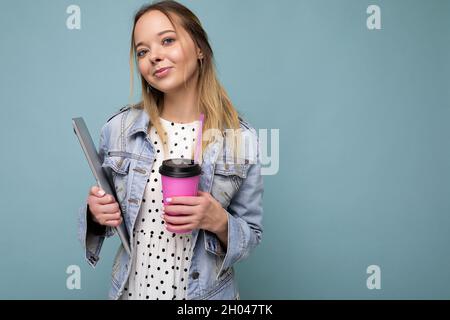 Image d'une joyeuse femme blonde européenne souriante et debout contre un mur bleu isolé avec un ordinateur portable argenté et un café à emporter dans les mains Banque D'Images