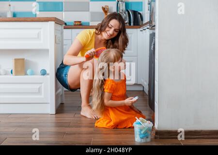 Jeune belle mère peignant sa fille avec peigne dans la cuisine tandis que l'enfant tire avec de la craie sur le réfrigérateur dans la cuisine intérieure confortable.Routine du matin Banque D'Images