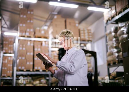 Un employé d'usine caucasien satisfait utilisant une tablette en se tenant dans l'entrepôt. Banque D'Images