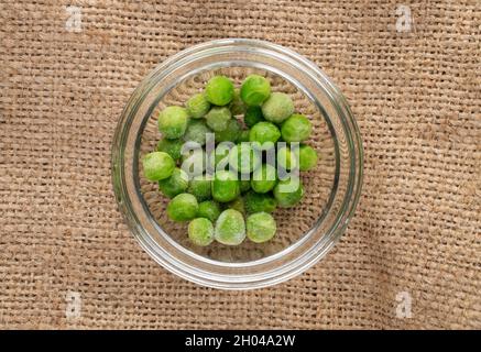 Plusieurs pois verts surgelés dans un plat en verre, gros plan, sur un tissu en jute, vue du dessus. Banque D'Images