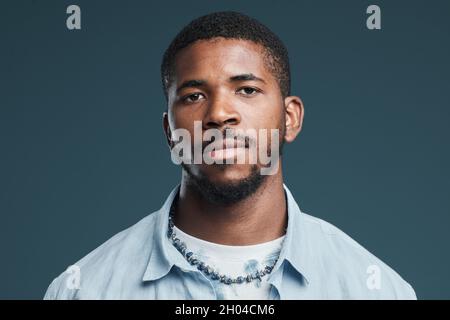 Portrait minimal de la tête et des épaules d'un beau homme afro-américain regardant l'appareil photo contre le bleu et portant des vêtements décontractés de rue Banque D'Images