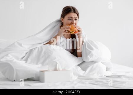 Bouleversée femme européenne millénaire affamée mangeant un gros hamburger, assis sur un lit blanc à l'intérieur de la chambre, a des problèmes nutritionnels, l'obésité, la dépendance alimentaire.ONU Banque D'Images