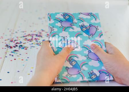 Mains d'une fille tenant un cahier avec licorne lama et arc-en-ciel sur fond blanc en bois avec confetti.Idée de table de bureau Girly.Retour à l'école con Banque D'Images