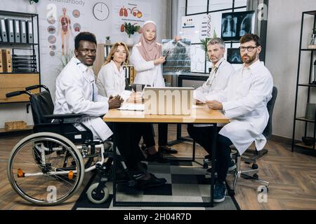 Groupe multiethnique de médecins regardant la caméra pendant la conférence au bureau de l'hôpital.Femme musulmane dans le hijab tenant une radiographie, homme afro-américain assis en fauteuil roulant.Concept d'inclusion. Banque D'Images