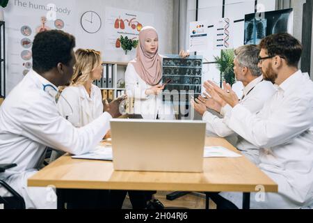 Une femme musulmane médecin en hijab discutant avec ses collègues multiraciaux d'un scanner à rayons X de patients graves.Cinq spécialistes médicaux à la recherche d'un mode de traitement de la santé lors d'une réunion au bureau. Banque D'Images