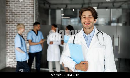 Beau jeune homme en vêtements de travail médecin assistant à la conférence médicale, tenant le tableau avec l'information, souriant à la caméra, groupe international de médecins Banque D'Images