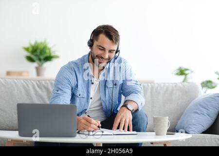 Un homme européen adulte souriant avec une barbe dans un casque parle au client et travaille à distance derrière un ordinateur portable dans le salon.Assistance clientèle, Banque D'Images