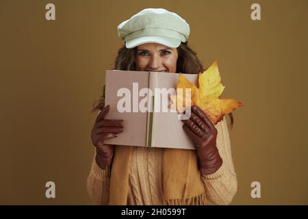 Bonjour septembre. Heureuse femme d'âge moyen en foulard avec feuille d'érable jaune d'automne, livre et gants en cuir sur fond marron. Banque D'Images